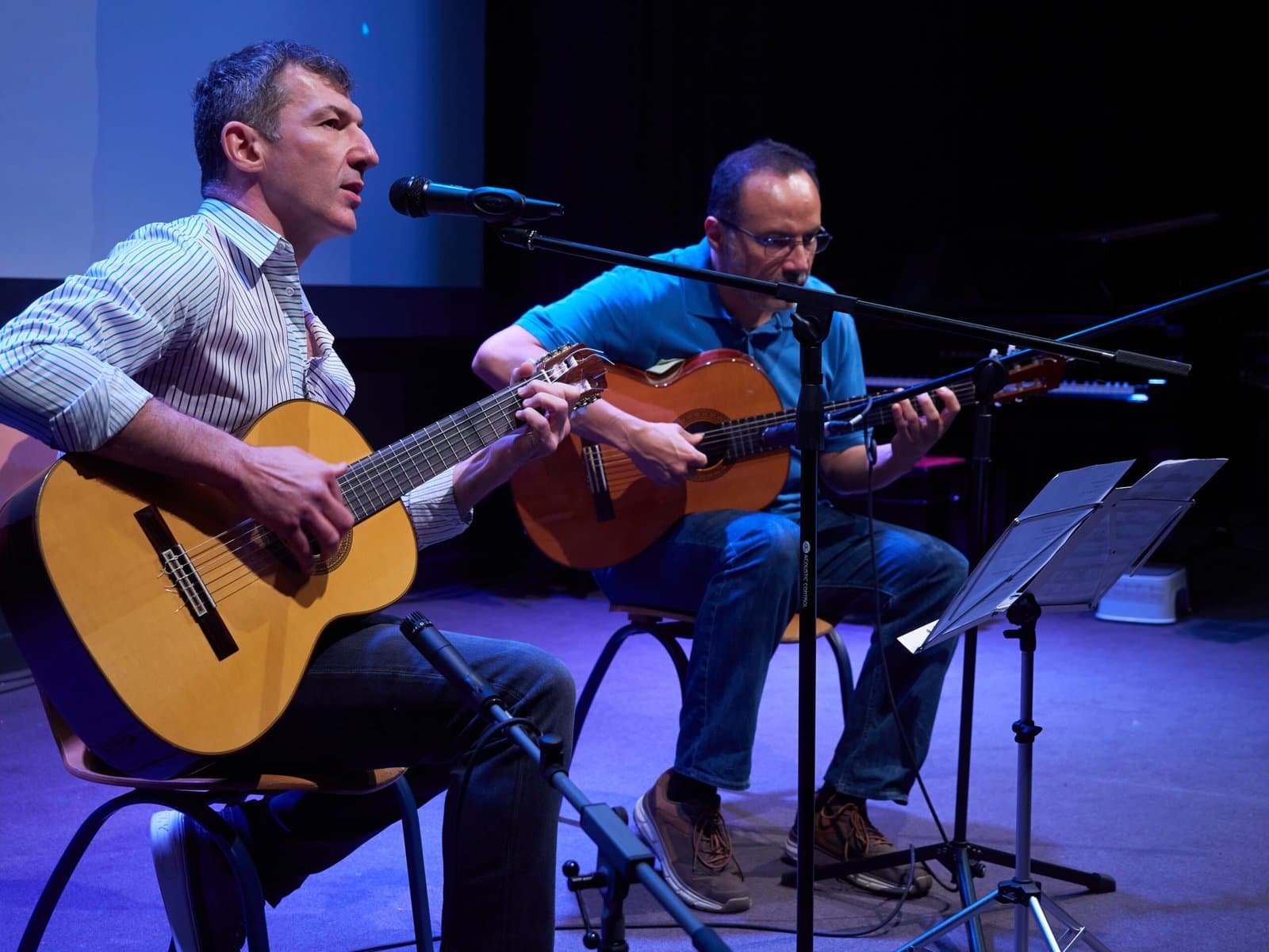 Dos músics tocant la guitarra en un escenari amb micròfons i partitures.