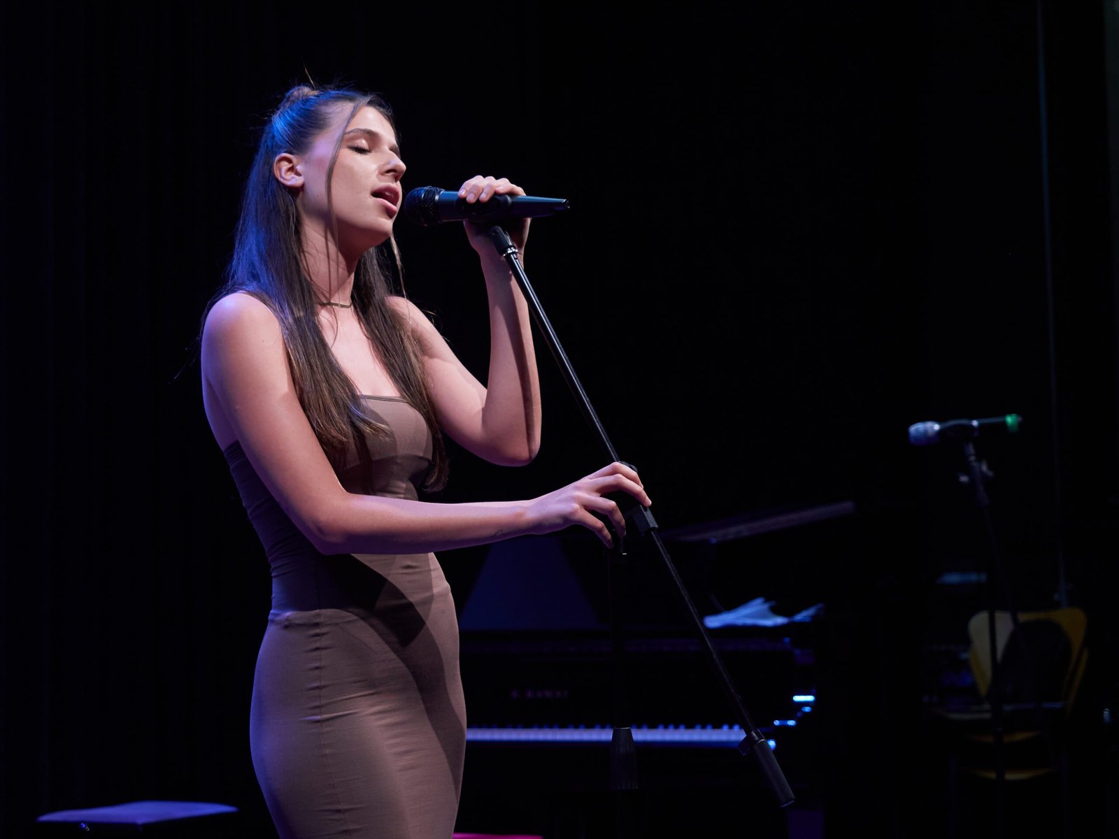 Joven mujer cantando en escenario con micrófono y piano al fondo.